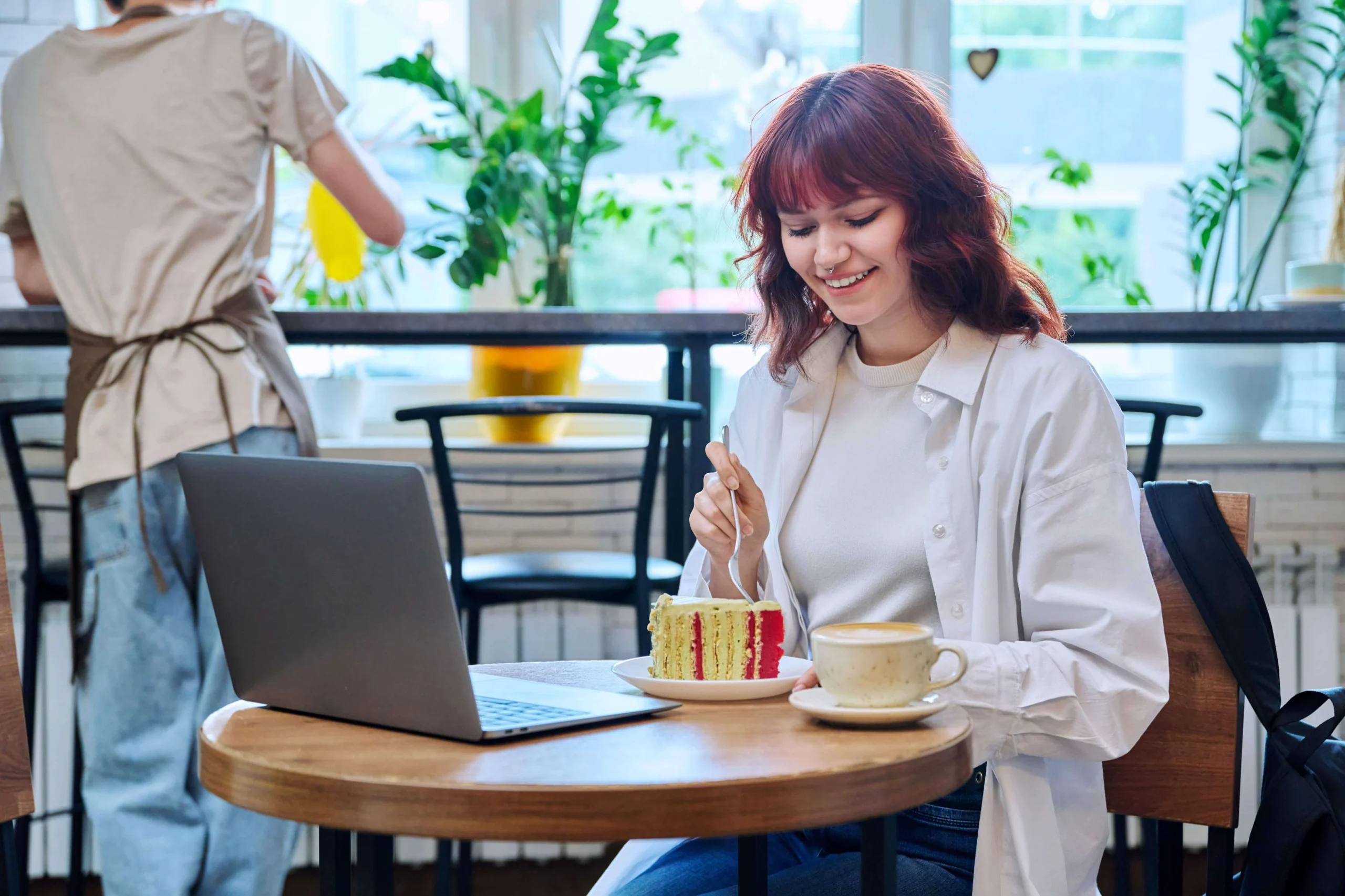 young woman enjoying coffee and cake while using a laptop in a café with free Wi-Fi.